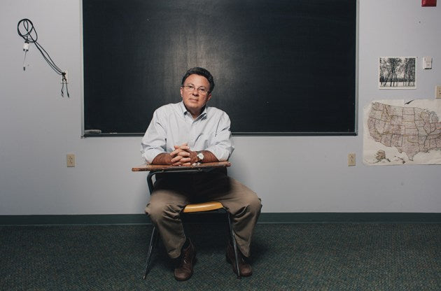 man sitting a child desk with chalkboard in the background