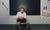 man sitting a child desk with chalkboard in the background