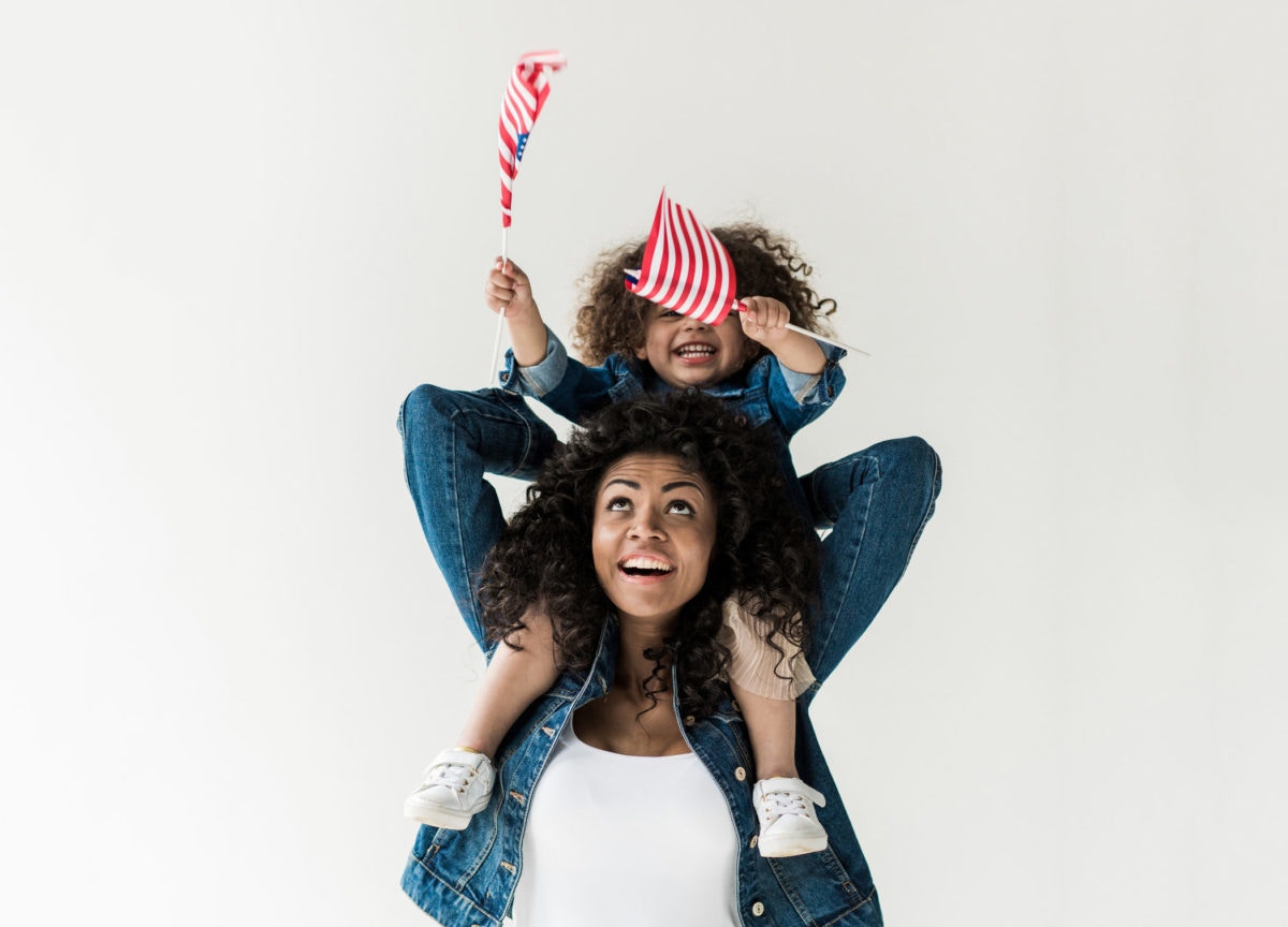 Little girl holding the flag of USA sits on her mother's shoulders