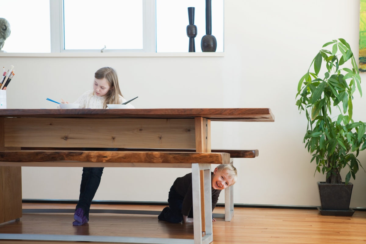 Girl is sitting on a table doing something while boy is sitting down side of a table by sneaking outside