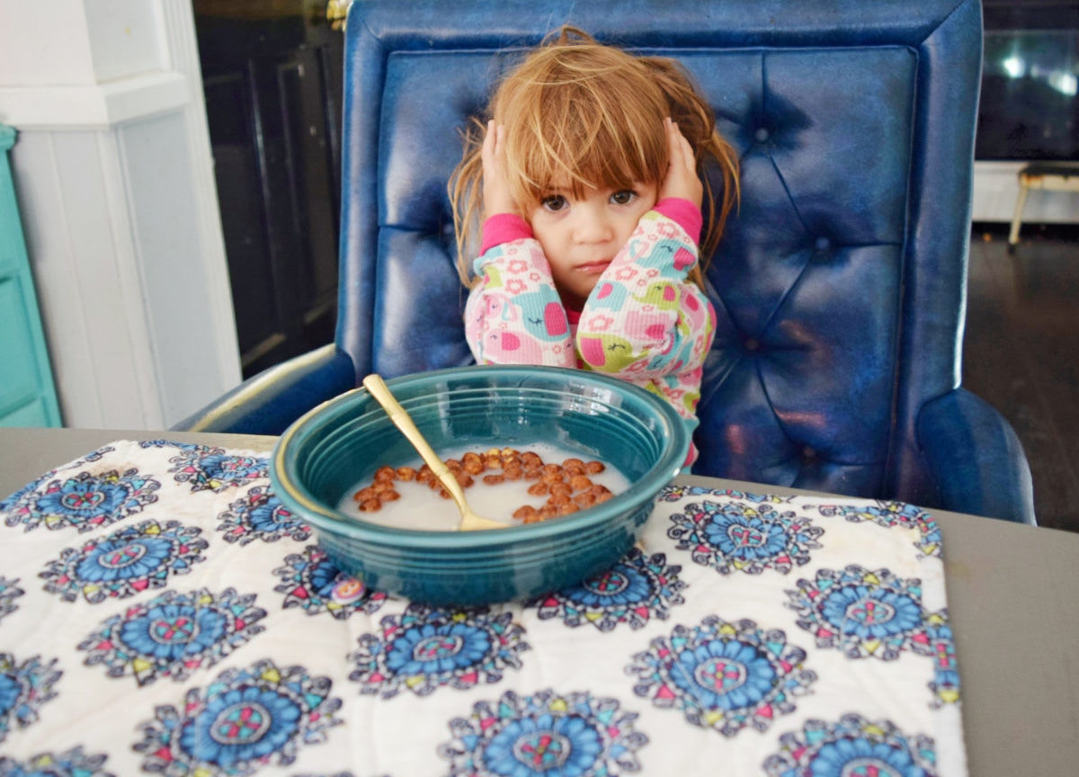 Little girl with annoyed expression sitting at a table with ears covered by her hands 