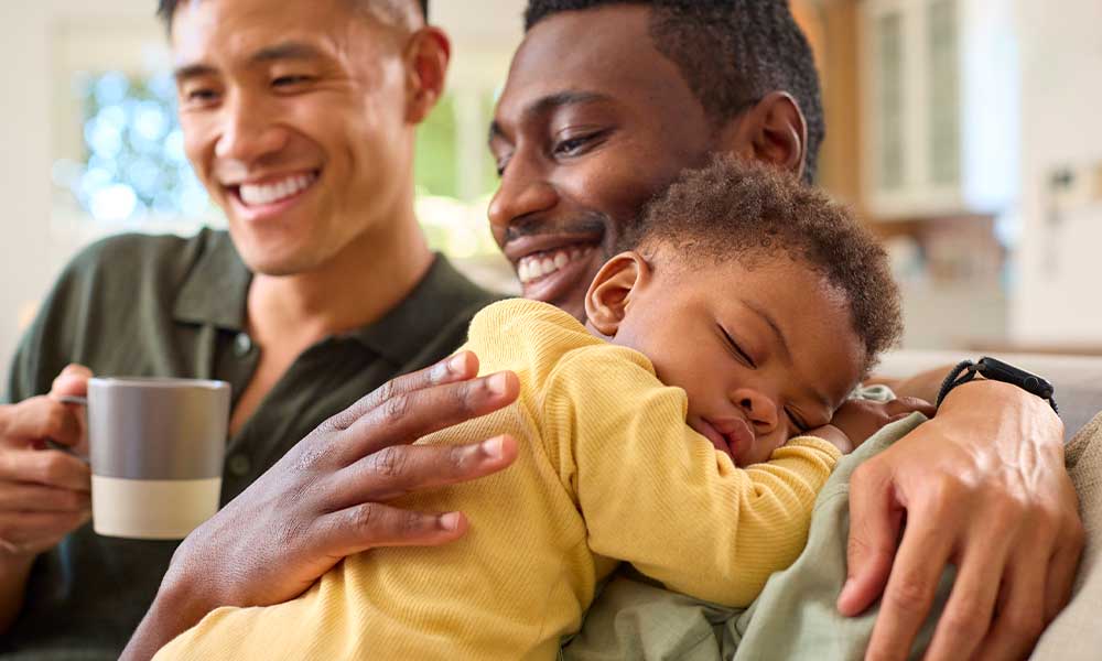 loving same sex male family cuddling, sleeping baby daughter on sofa